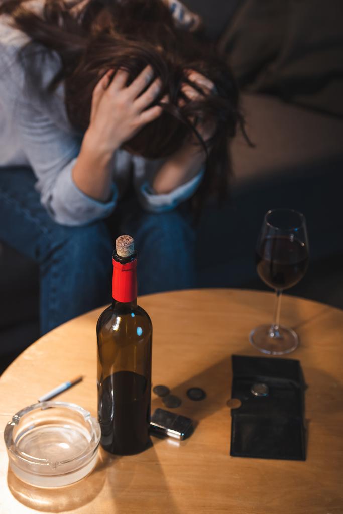 alcoholverslaafde vrouw met gebogen hoofd in de buurt van fles rode wijn, munten en lege portemonnee, wazige achtergrond     - Foto, afbeelding