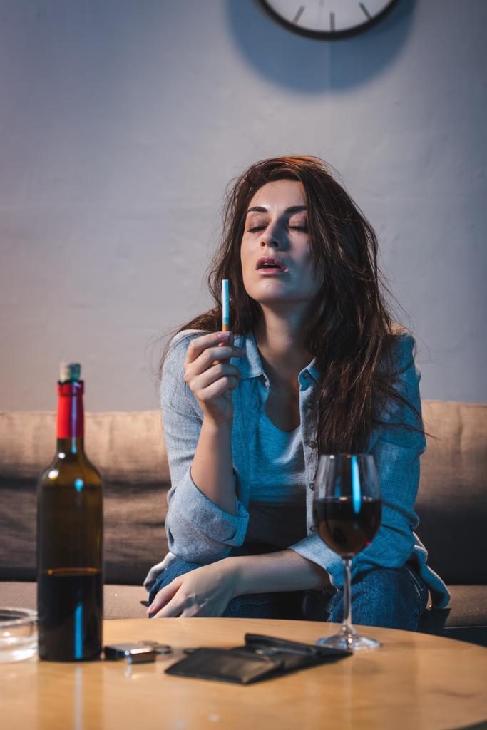 テーブルの上に赤ワインと空の財布の近くに座っている間にタバコを持っている酔った女性 - 写真・画像