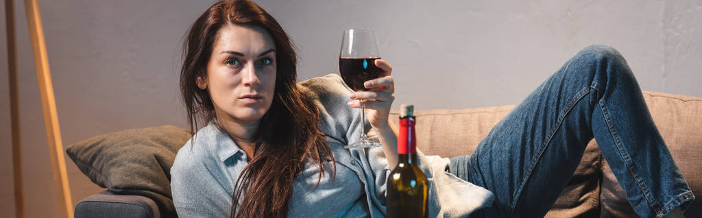 μεθυσμένη, απογοητευμένη γυναίκα κοιτάζει την κάμερα ενώ ξαπλώνει με ένα ποτήρι κόκκινο κρασί, πανό - Φωτογραφία, εικόνα