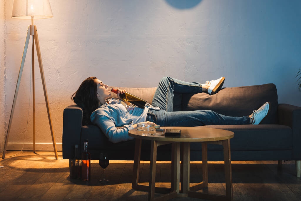 vista lateral de la mujer deprimida bebiendo vino de la botella mientras está acostada en el sofá - Foto, imagen