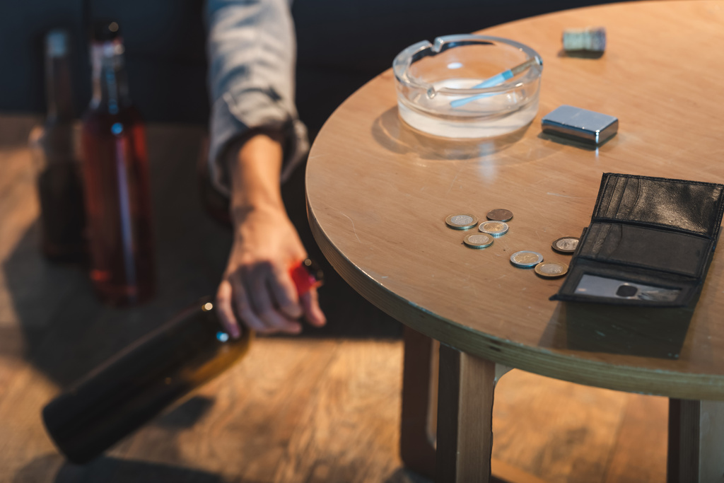 gedeeltelijke weergave van vrouwelijke hand met fles alcohol in de buurt van munten en lege portemonnee op tafel, wazige achtergrond  - Foto, afbeelding