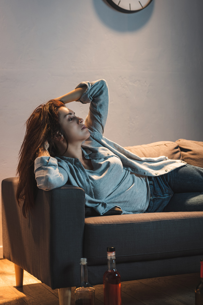 καταθλιπτική, εθισμένη στο αλκοόλ γυναίκα ξαπλωμένη στον καναπέ κοντά σε μπουκάλια στο πάτωμα - Φωτογραφία, εικόνα