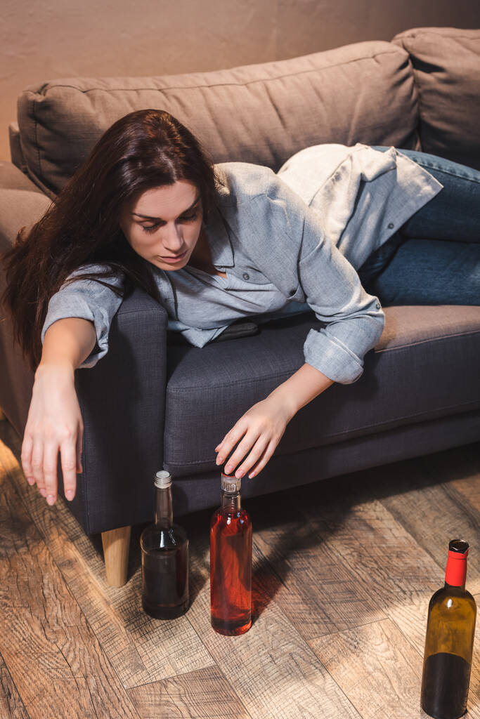 μεθυσμένη γυναίκα ξαπλωμένη στον καναπέ κοντά σε μπουκάλια αλκοόλ στο πάτωμα.  - Φωτογραφία, εικόνα