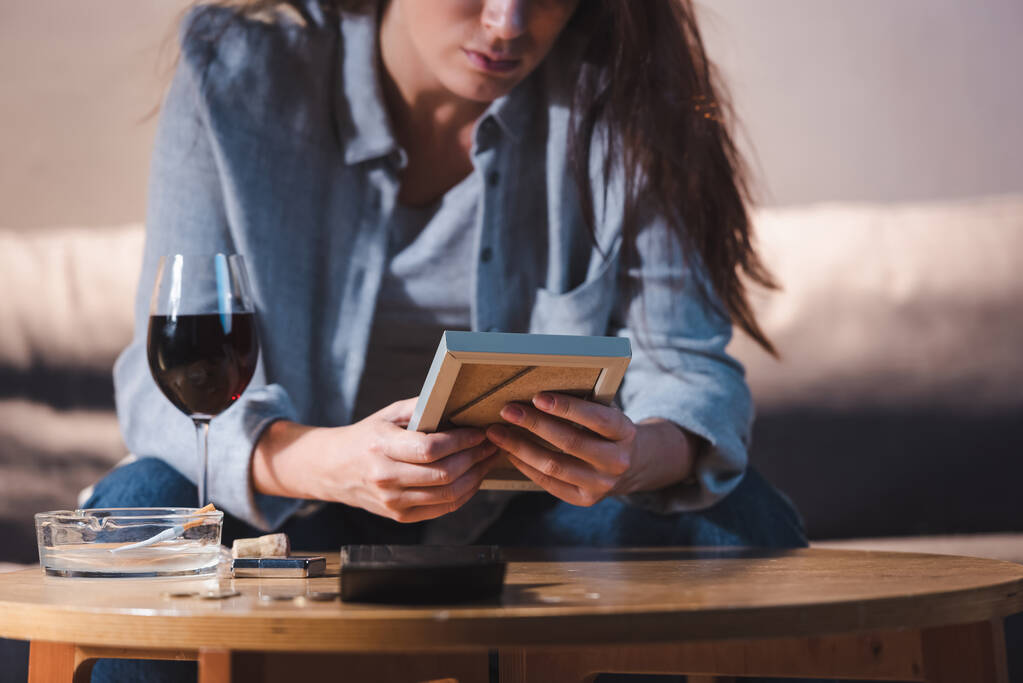 cropped view of depressed woman holding photo frame near glass of red wine, blurred background - Photo, Image