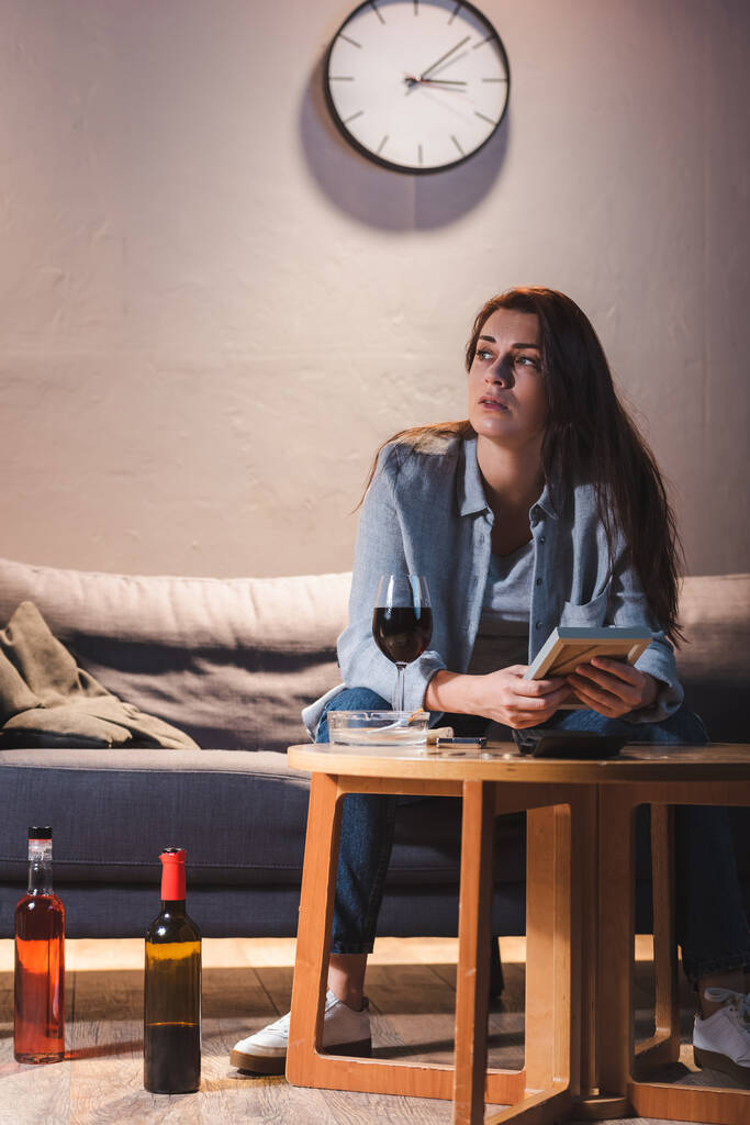 καταθλιπτική γυναίκα κοιτάζει μακριά, ενώ κάθεται κοντά σε μπουκάλια αλκοόλ και κρατώντας κορνίζα φωτογραφία - Φωτογραφία, εικόνα