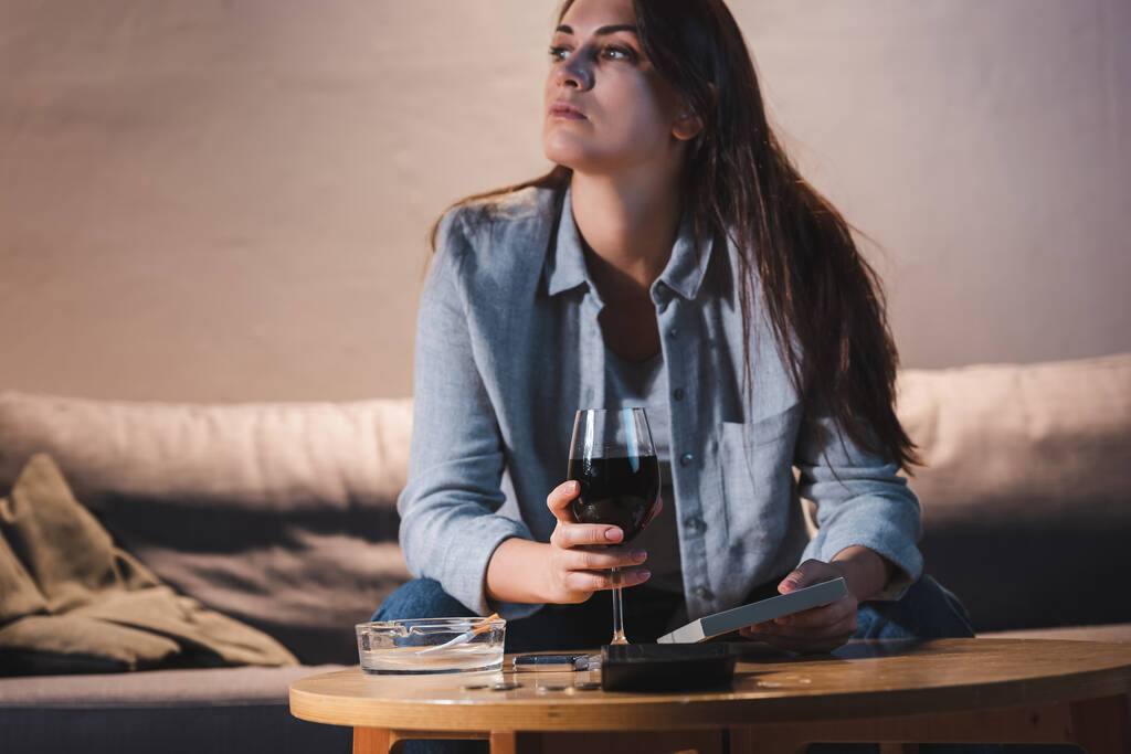 frustrovaná žena závislá na alkoholu se dívá stranou, zatímco sedí s fotorámečkem a sklenicí vína     - Fotografie, Obrázek