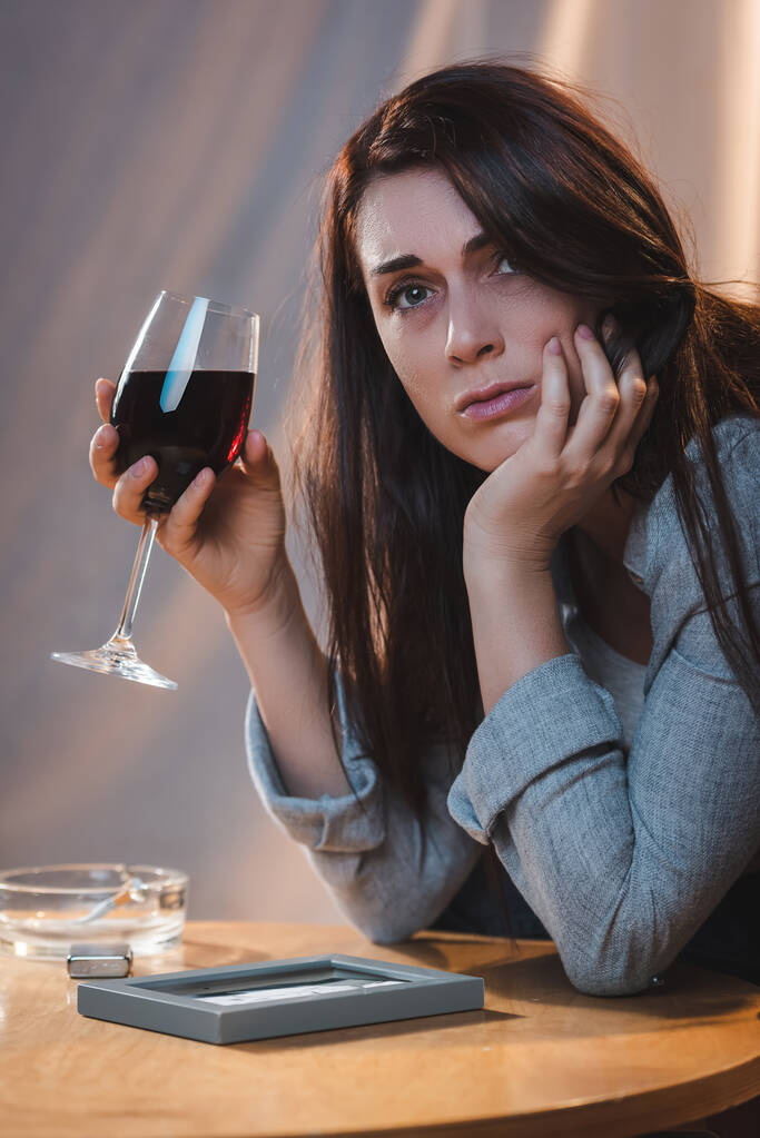 απογοητευμένη γυναίκα κοιτάζοντας μακριά, ενώ κάθεται κοντά στο πλαίσιο φωτογραφία με ένα ποτήρι κόκκινο κρασί - Φωτογραφία, εικόνα