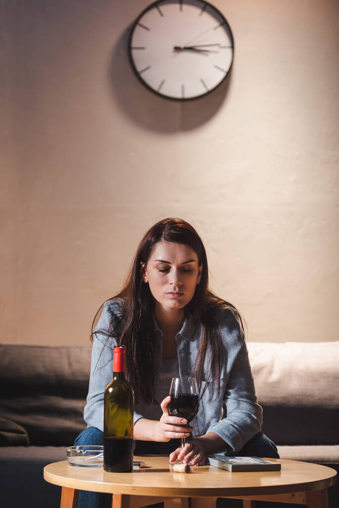 Şişenin yanında otururken elinde kırmızı şarap kadehi tutan depresif kadın ve masanın üzerindeki fotoğraf çerçevesi. - Fotoğraf, Görsel