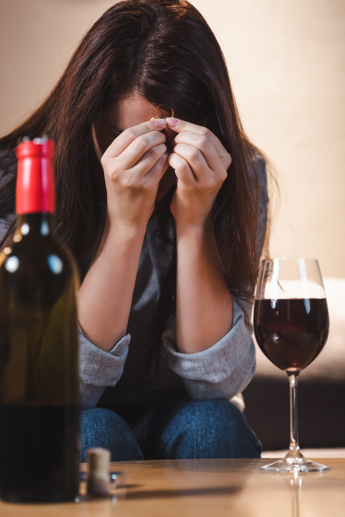 mujer deprimida y adicta que oscurece la cara con las manos mientras sostiene el anillo de bodas cerca del vino tinto en un primer plano borroso    - Foto, imagen