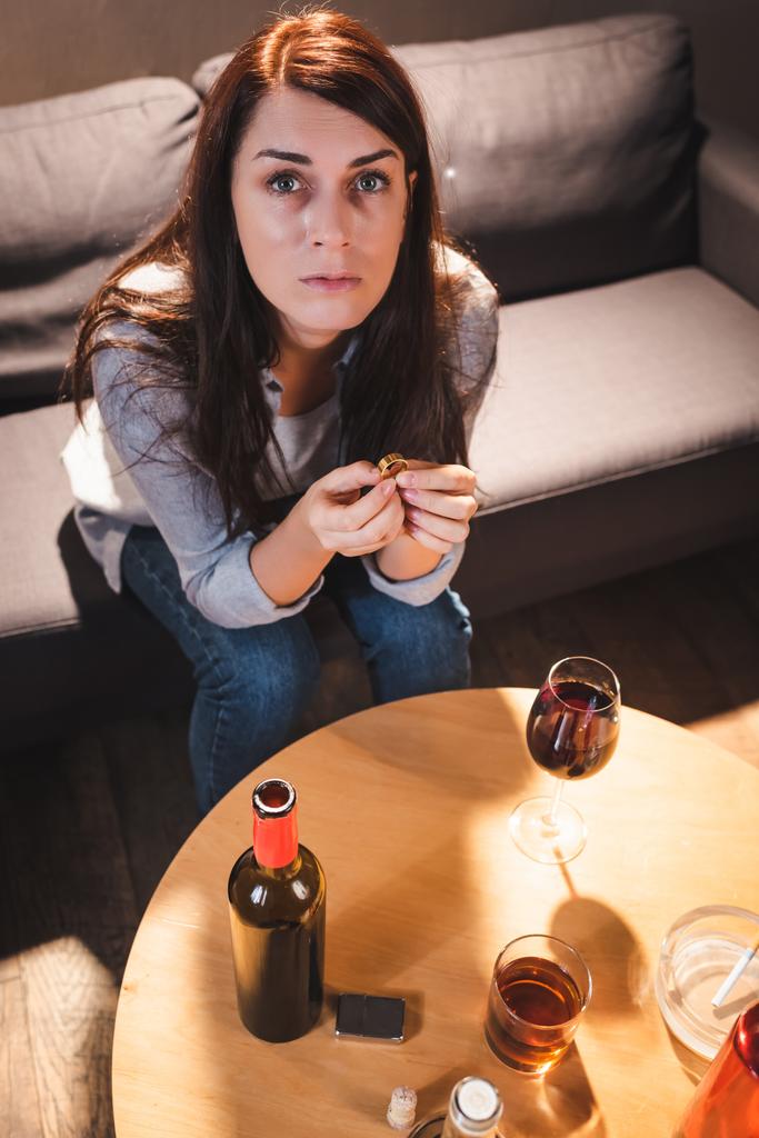 wysoki kąt widzenia depresji kobieta patrząc w aparat podczas siedzenia w pobliżu napojów alkoholowych i trzymając obrączkę - Zdjęcie, obraz