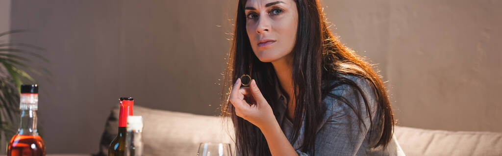 depresivní žena závislá na alkoholu při pohledu do kamery, zatímco drží snubní prsten blízko lahví na rozmazaném popředí, banner - Fotografie, Obrázek