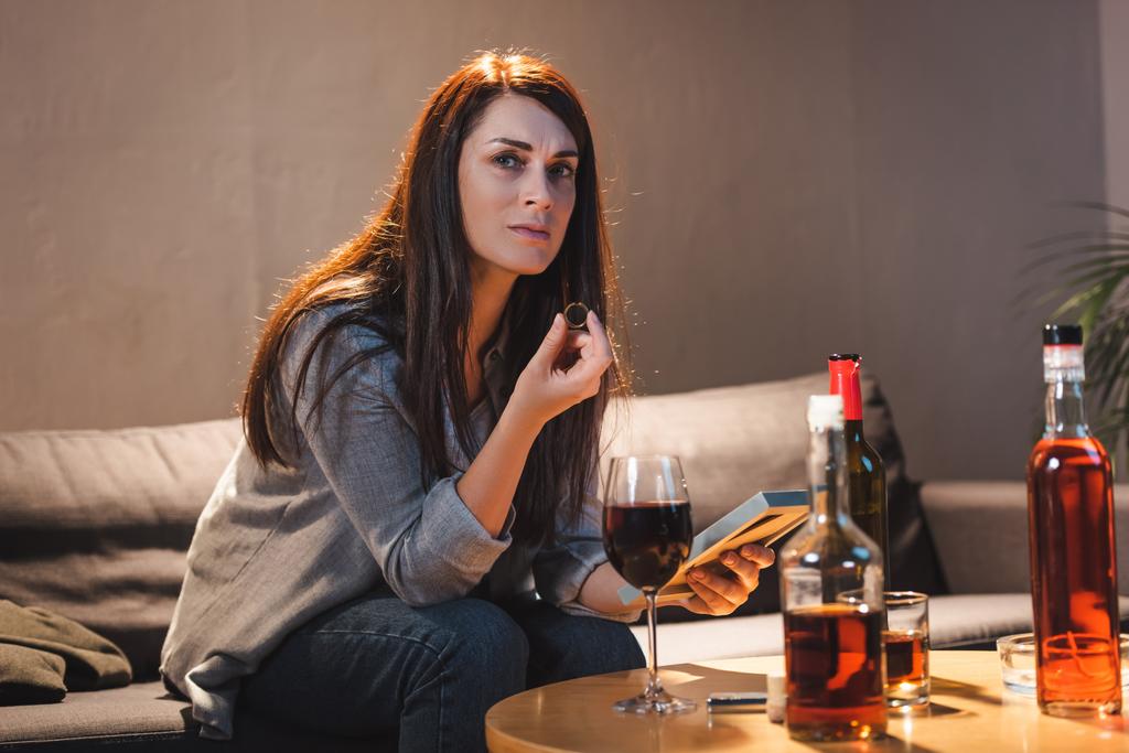 Frustrierte Frau blickt in die Kamera, während sie Ehering und Fotorahmen in der Nähe von Alkoholgetränken auf dem Tisch hält - Foto, Bild