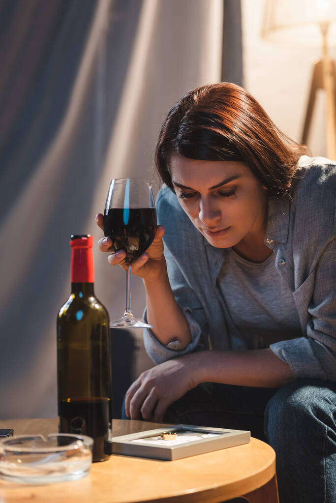 zdenerwowana, samotna kobieta patrząca na ramkę ze zdjęciem trzymając kieliszek czerwonego wina - Zdjęcie, obraz