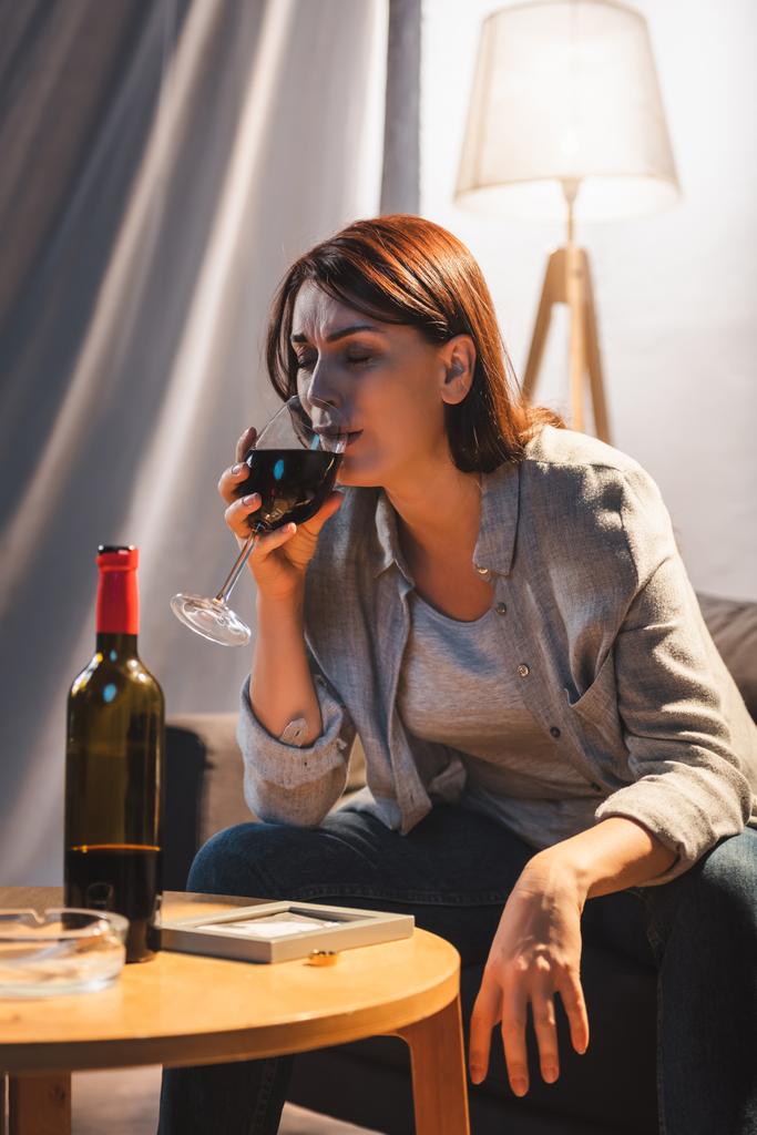 mujer frustrada llorando y bebiendo vino mientras se sienta cerca de marco de fotos y anillo de boda en la mesa  - Foto, Imagen