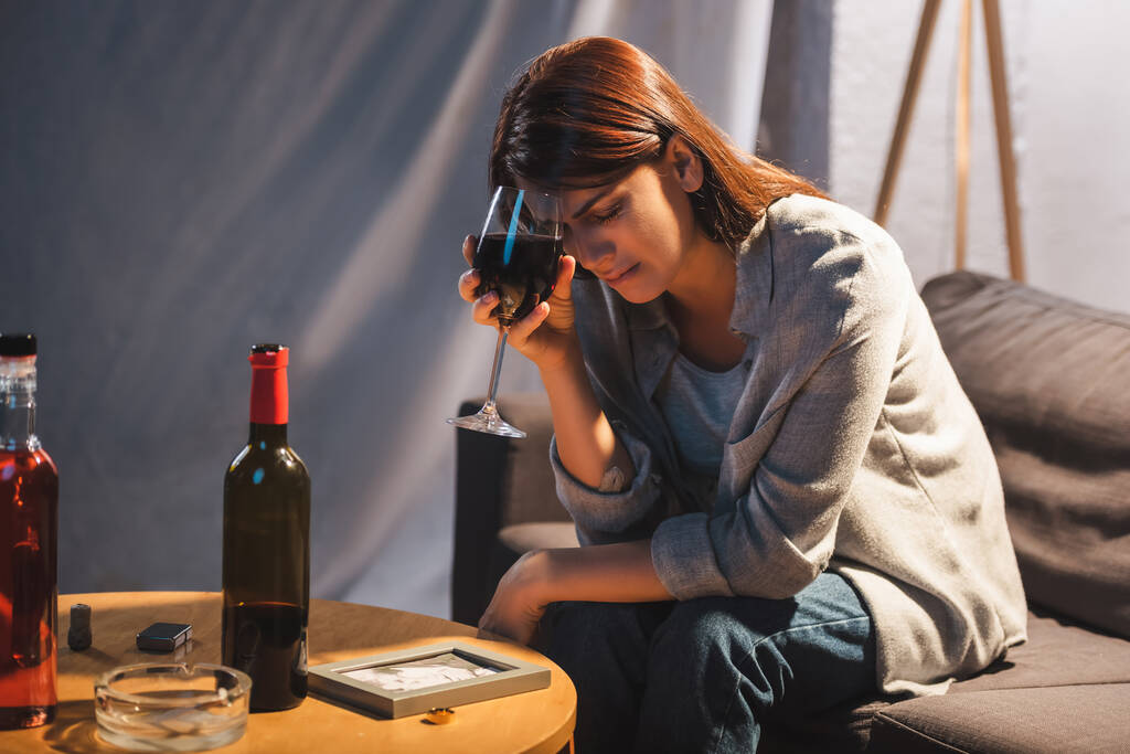 depresivní žena sedící se zavřenýma očima a sklenicí vína poblíž fotorámečku a snubního prstenu na stole - Fotografie, Obrázek