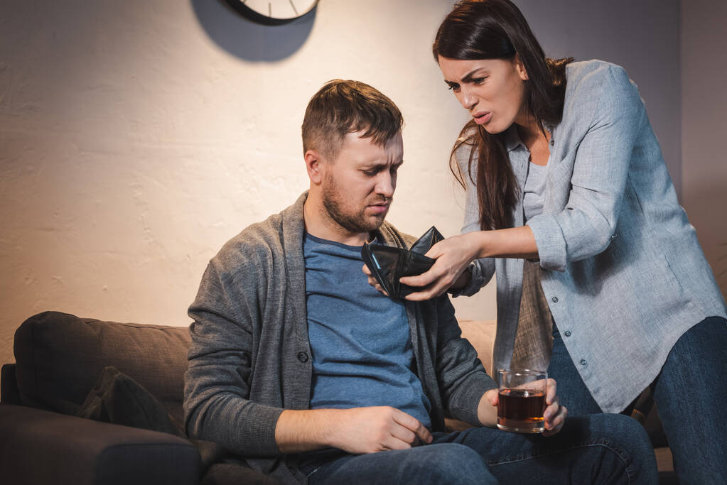 geïrriteerde vrouw tonen lege portemonnee naar dronken man met glas whisky - Foto, afbeelding