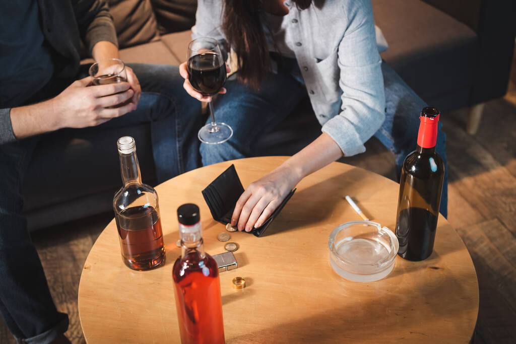 vista recortada de pareja sosteniendo vasos de alcohol cerca de botellas, monedas y billetera vacía en la mesa - Foto, Imagen