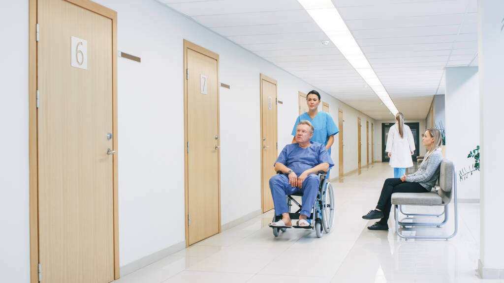 In the Hospital Hallway, Nurse Pushes Senior Man in the Wheelchair, Patients wait for their Doctor, Busy Professional Personnel Walking by. Clean, New Hospital with Professional Medical Staff. - Photo, Image