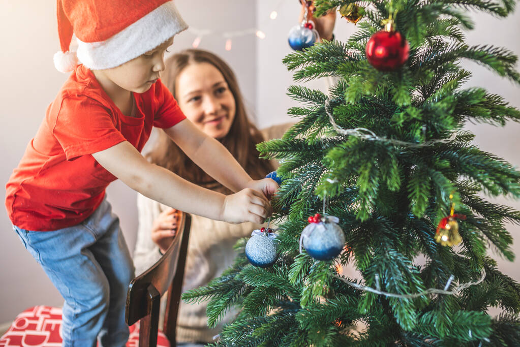 Matka se svým synem dítě zdobí vánoční stromek s hračkami a vánoční koule. Šťastný rodinný čas na Silvestra doma - Fotografie, Obrázek