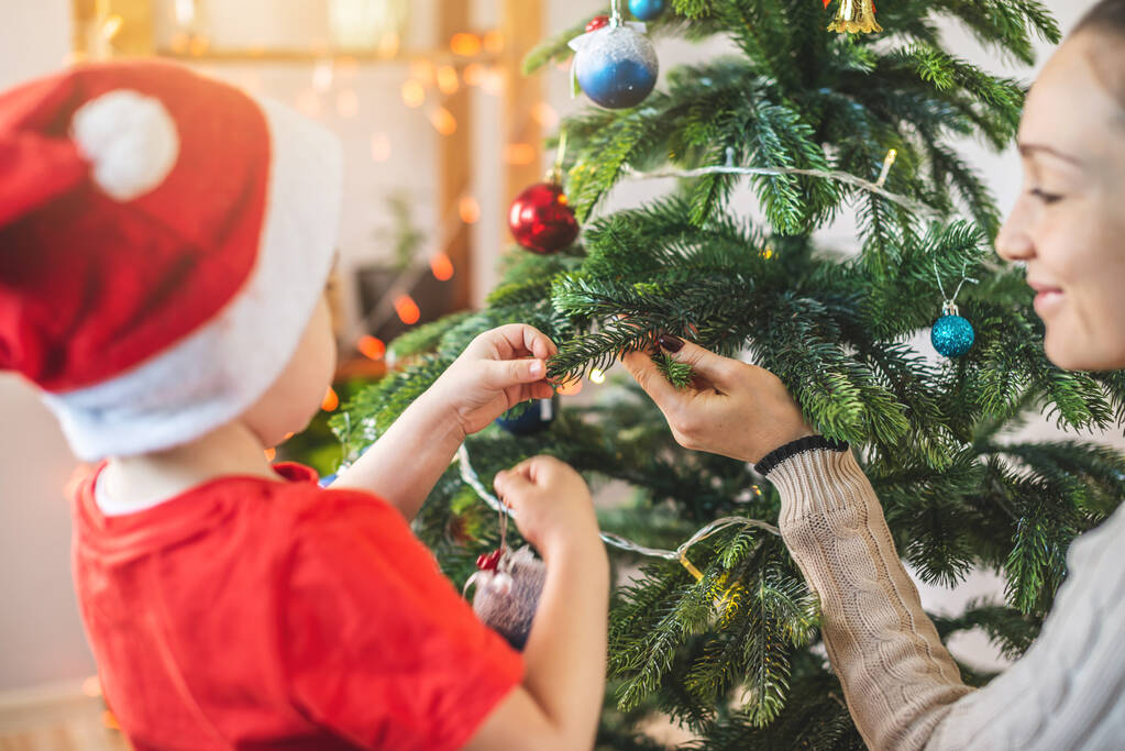 Anya a gyermekével fia díszíti a karácsonyfát játékokkal és karácsonyi labdákkal. Boldog családi estét itthon szilveszterkor. - Fotó, kép