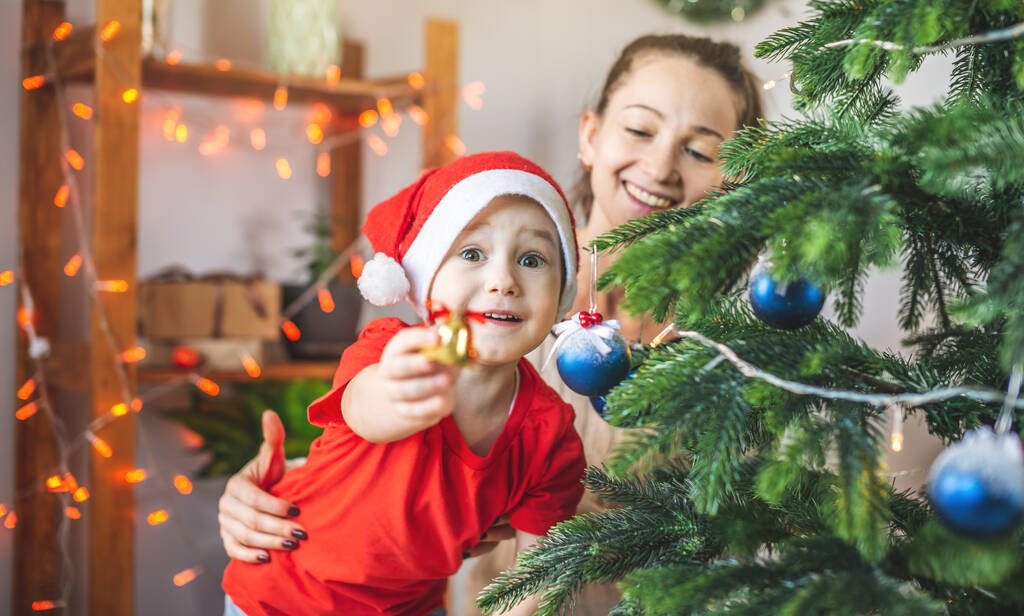 Mom is helping her child son decorate the Christmas tree with toys and Christmas balls. The concept of a happy loving family on new year's eve - Photo, Image