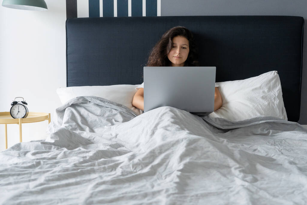 Gyönyörű barna lány az ágyban egy laptoppal. Reggeltől késő estig dolgozom. Elektronikus kütyü lefekvés előtt vagy reggel - Fotó, kép