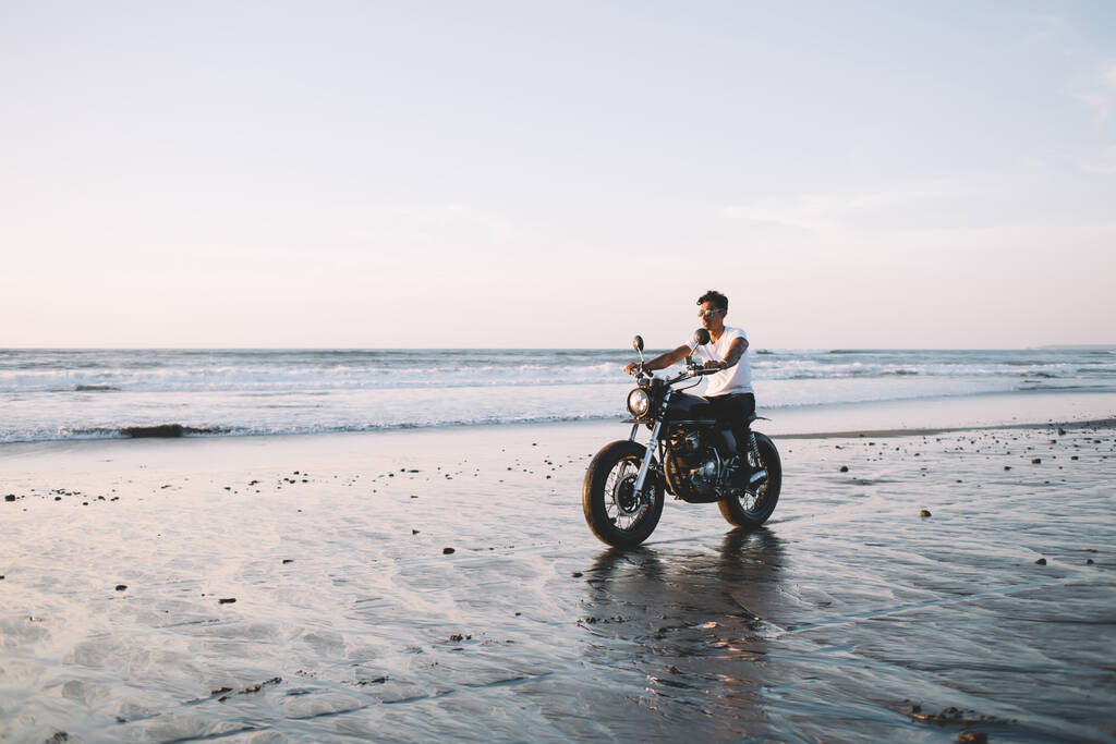 Jovem piloto étnico masculino em casual desgaste leve e óculos descansando enquanto andava de moto ao longo da costa arenosa deserta ao pôr do sol - Foto, Imagem