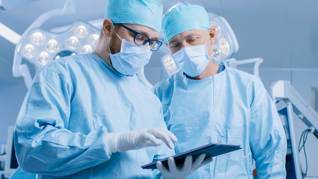 Dwóch profesjonalnych chirurgów podczas stania w nowoczesnej sali operacyjnej szpitala korzysta z komputera z tabletem cyfrowym. - Zdjęcie, obraz