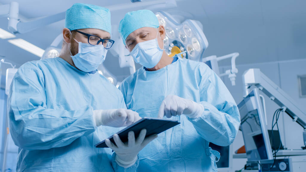 Dos cirujanos profesionales utilizan la computadora digital de la tableta mientras que están parados en la sala de operaciones moderna del hospital. - Foto, Imagen