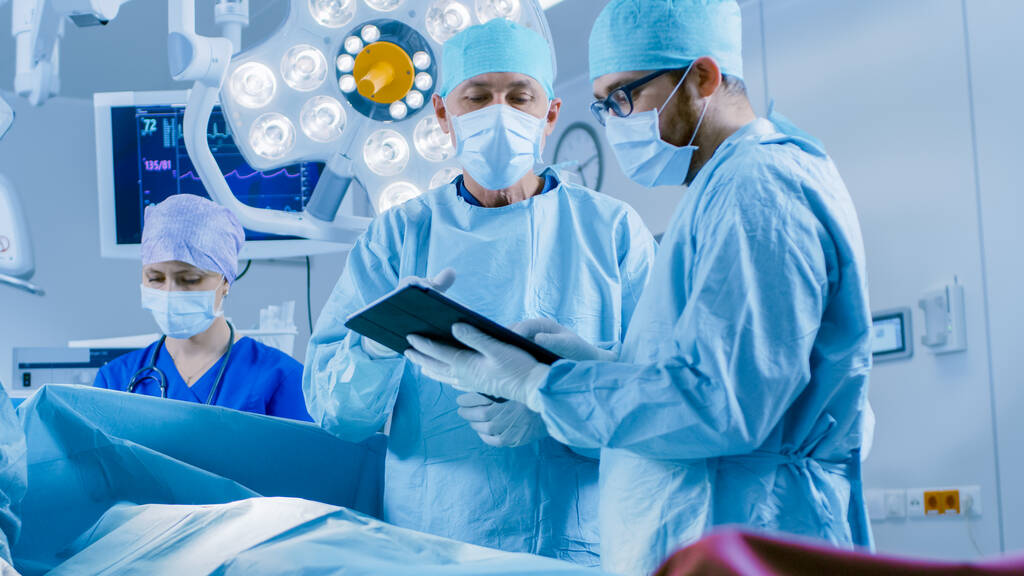 Chirurghi professionisti e assistenti Parlare e utilizzare il computer tablet digitale durante l'intervento chirurgico. Lavorano nella moderna sala operatoria dell'ospedale. - Foto, immagini