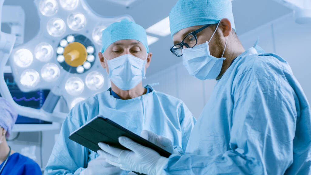 Due chirurghi professionisti usano il computer digitale della compressa mentre si levano in piedi nella sala operatoria moderna dell'ospedale. - Foto, immagini