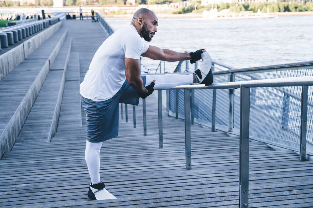 Πλευρική άποψη της προνοητικής bodybuilder σε αθλητικά ρούχα και γάντια άσκηση το πρωί προθέρμανση πόδια σε μεταλλικό φράχτη σκάλες κοιτάζοντας μακριά στο φόντο της προκυμαίας - Φωτογραφία, εικόνα
