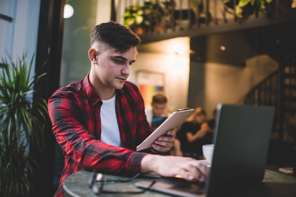 Od dołu zamyślony młody mężczyzna w nieformalnej koszuli wyszukiwanie informacji z schowka na laptopie podczas siedzenia przy stole w kawiarni - Zdjęcie, obraz