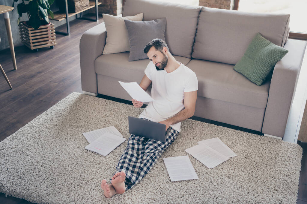 Nice attractive focused brunet guy sitting on carpet bare foot working on laptop project start-up analyzing documents at modern loft industrial interior house apartment indoors - Photo, Image