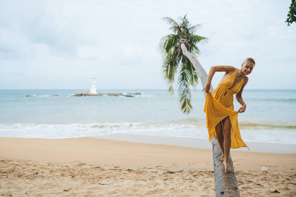 Счастливая молодая женщина в желтом платье идет вдоль ствола пальмы на пляже - Фото, изображение