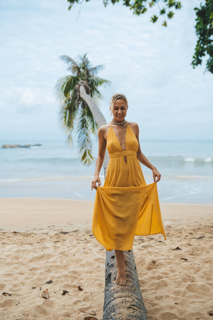 Jovem feliz em vestido amarelo caminhando ao longo do tronco de uma palmeira na praia - Foto, Imagem