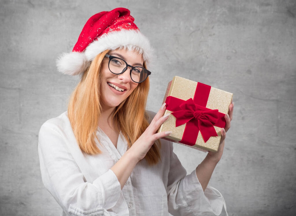 Νεαρή όμορφη ξανθιά γυναίκα φοράει Χριστούγεννα Σάντα Claus καπέλο - καπέλο και κρατώντας κόκκινο κουτί δώρου - παρουσιάζουν με κορδέλα, απομονώνονται σε γκρι φόντο. Φωτογραφία στούντιο - Φωτογραφία, εικόνα