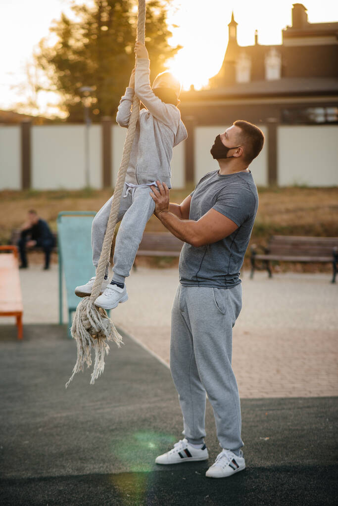 A father helps his son climb a rope on a sports field in masks during sunset. Healthy parenting and healthy lifestyle - Photo, Image