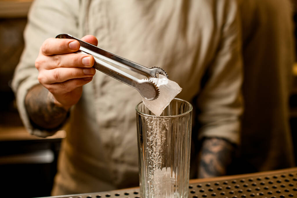 αρσενικό μπάρμαν κρατώντας λαβίδες στο χέρι με παγάκι πάνω από γυαλί στο μπαρ - Φωτογραφία, εικόνα