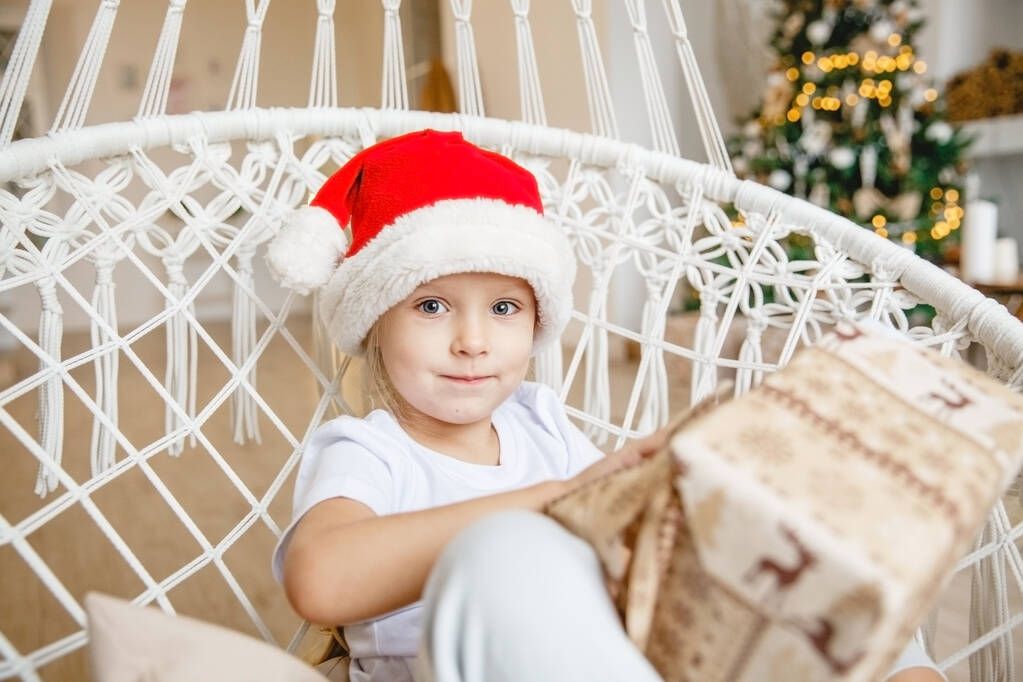 Ένα τετράχρονο κορίτσι με καπέλο Αϊ Βασίλη κάθεται σε μια ψάθινη καρέκλα στο φόντο ενός χριστουγεννιάτικου δέντρου και ξετυλίγει ένα δώρο. - Φωτογραφία, εικόνα
