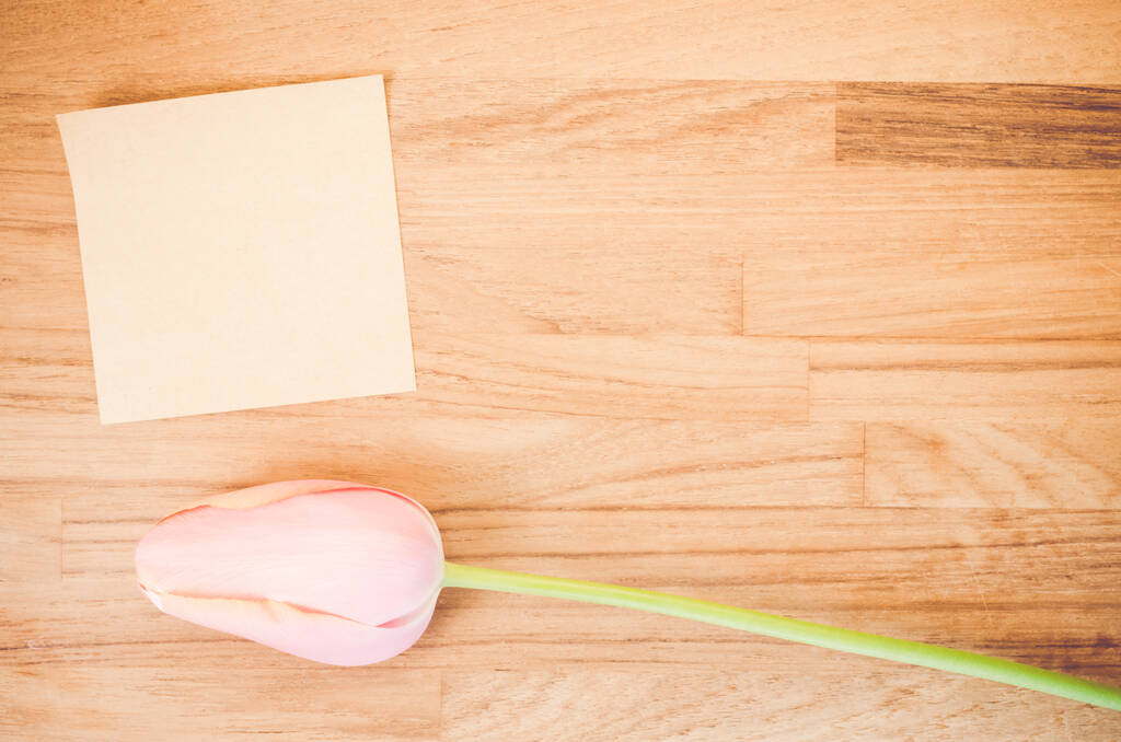 Un plano superior de una flor de tulipán rosa y una nota adhesiva en blanco en una mesa de madera con espacio para su texto - Foto, Imagen