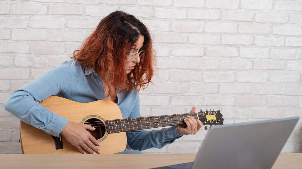 Běloška se učí hrát na kytaru online. Dívka sedí na dálku v karanténě a studuje hudbu - Fotografie, Obrázek