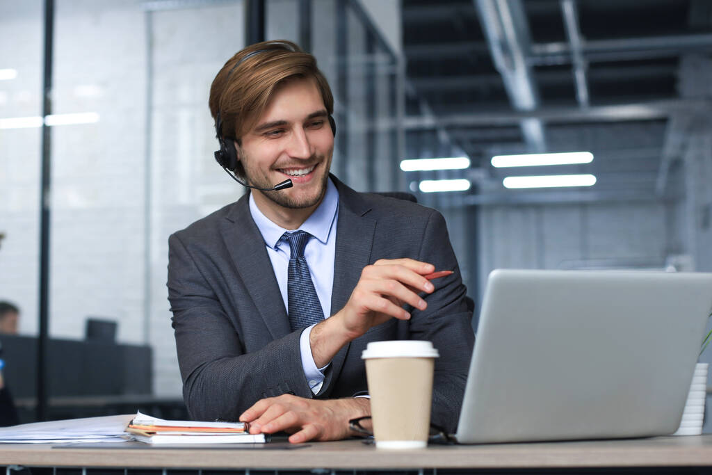 Kulaklıklı, gülümseyen bir erkek çağrı merkezi operatörü, modern ofiste oturuyor, bir dizüstü bilgisayardaki çevrimiçi bilgilere danışıyor, müşteriye yardımcı olmak için bir dosyada bilgilere bakıyor. - Fotoğraf, Görsel