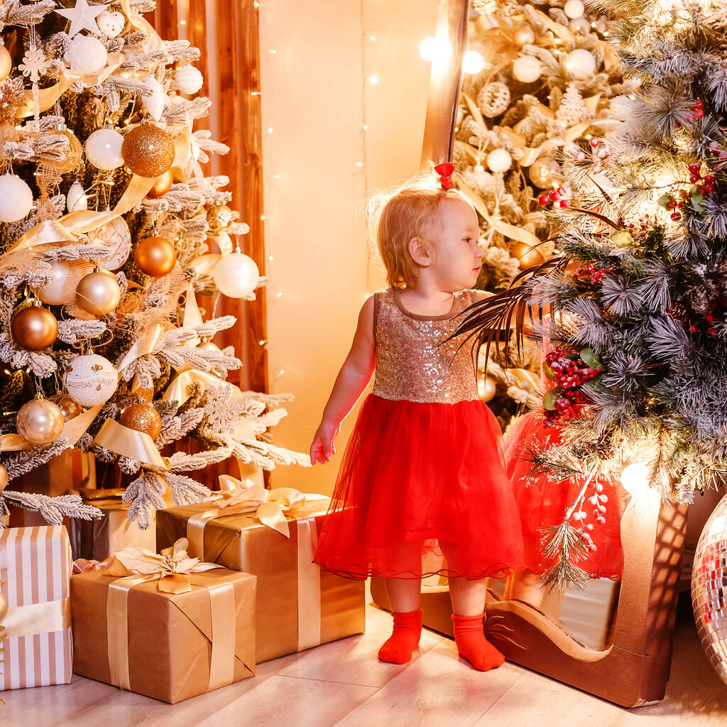 Маленька дівчинка в червоній сукні дивиться у дзеркало в різдвяній залі
 - Фото, зображення
