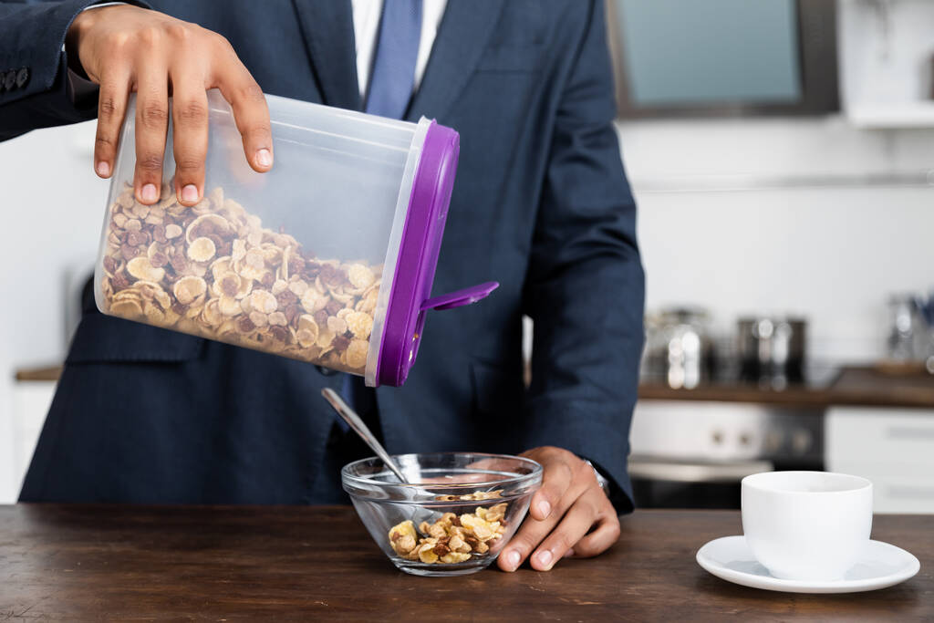gedeeltelijk zicht op Afrikaans-Amerikaanse zakenman die container met cornflakes in de buurt van kom houdt - Foto, afbeelding