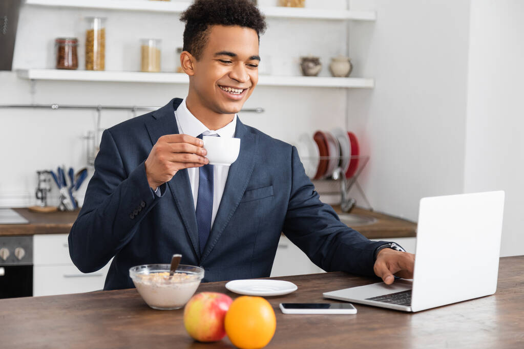 щасливий афроамериканський фрілансер у костюмі з ноутбуком під час сніданку
  - Фото, зображення