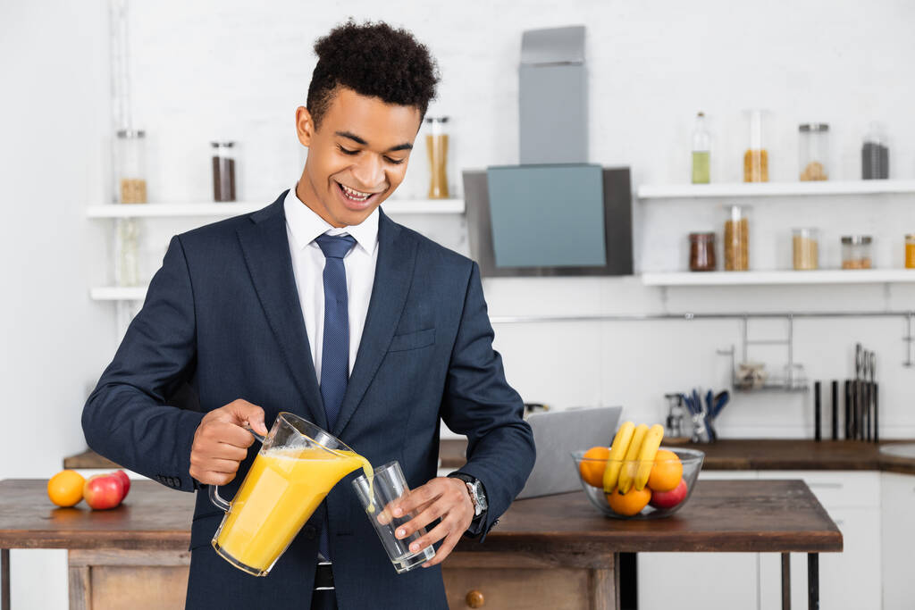 heureux homme d'affaires afro-américain tenant cruche et verser du jus d'orange dans le verre  - Photo, image