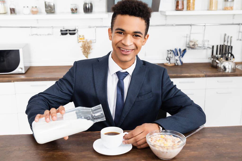 ευτυχισμένος Αφρικανός Αμερικανός επιχειρηματίας ρίχνει γάλα σε καφέ κοντά στο μπολ με νιφάδες καλαμποκιού  - Φωτογραφία, εικόνα