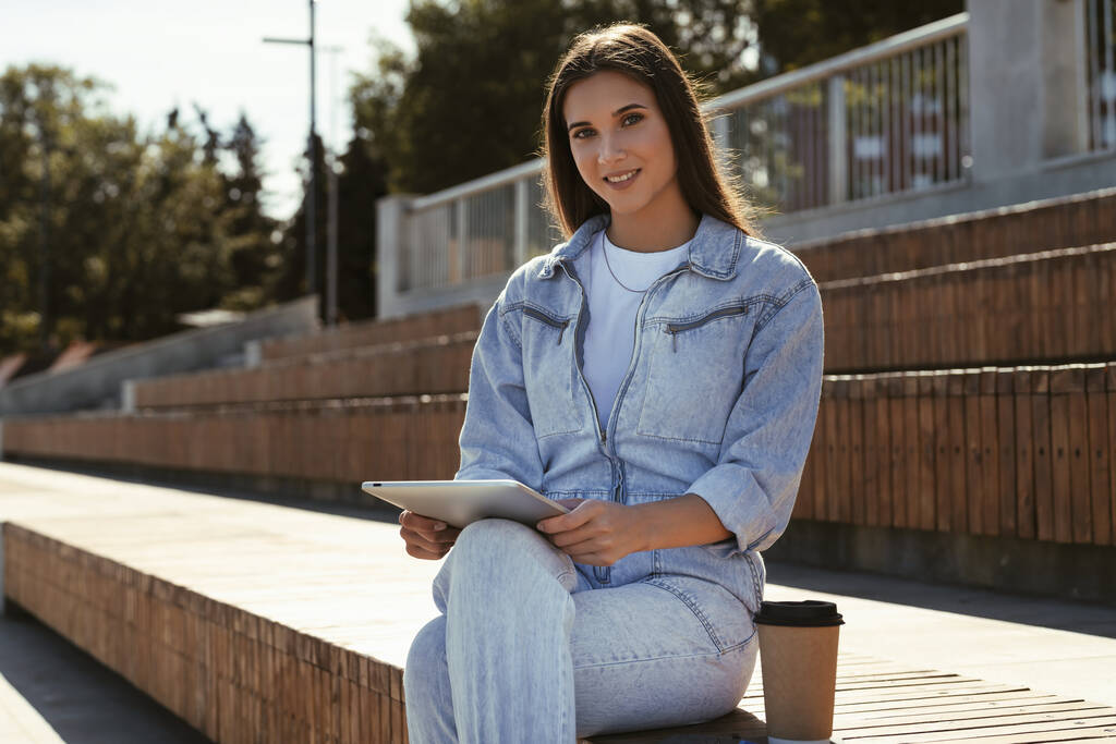 Приваблива дівчина в джинсовому одязі сидить на лавці парку, тримає розумний планшет в руках. Портрет красивої жінки
 - Фото, зображення