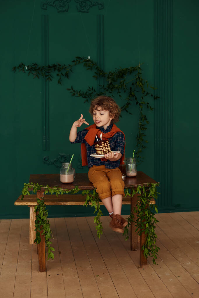 Cute kręcone mały chłopiec świętuje swoje urodziny z pyszne naleśniki wakacje. Dziecko w stożkowym kapeluszu świętuje na przyjęciu domowym w stylowym pokoju z zielonymi ścianami ozdobionymi liśćmi. - Zdjęcie, obraz